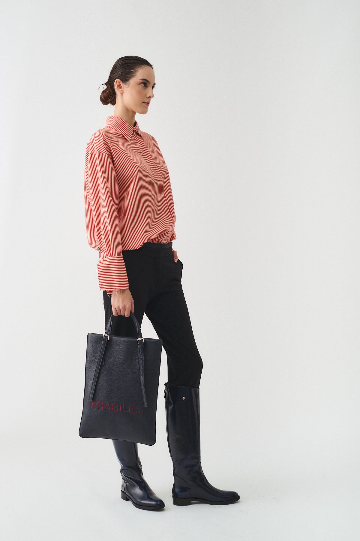 Modella indossa la CAMICIA ISOTTA IN SETA CON BOTTONI GIOIELLO, pantaloni neri e stivali alti, mentre tiene una borsa nera.