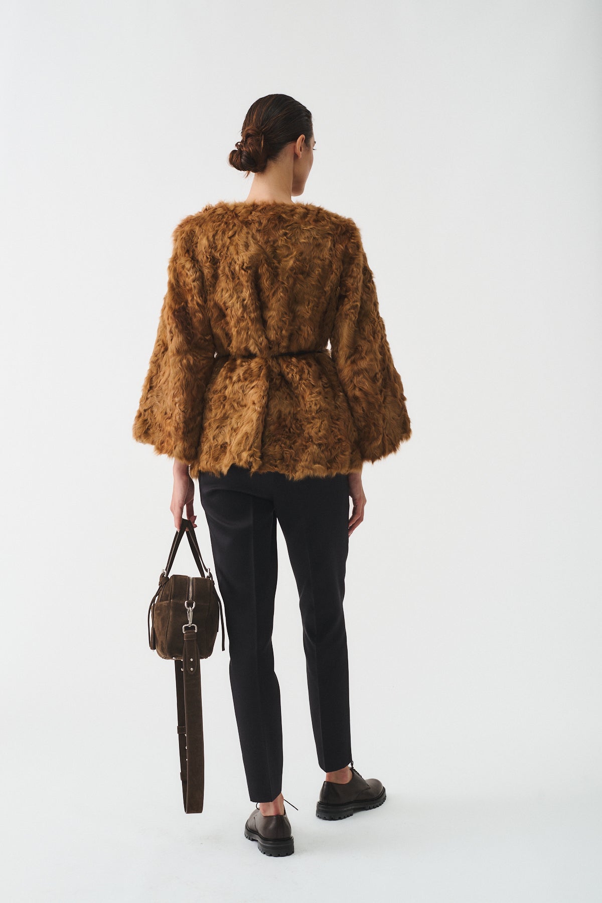 Back view of a model in a brown fur caban with a belt, paired with black pants and holding a stylish bag.