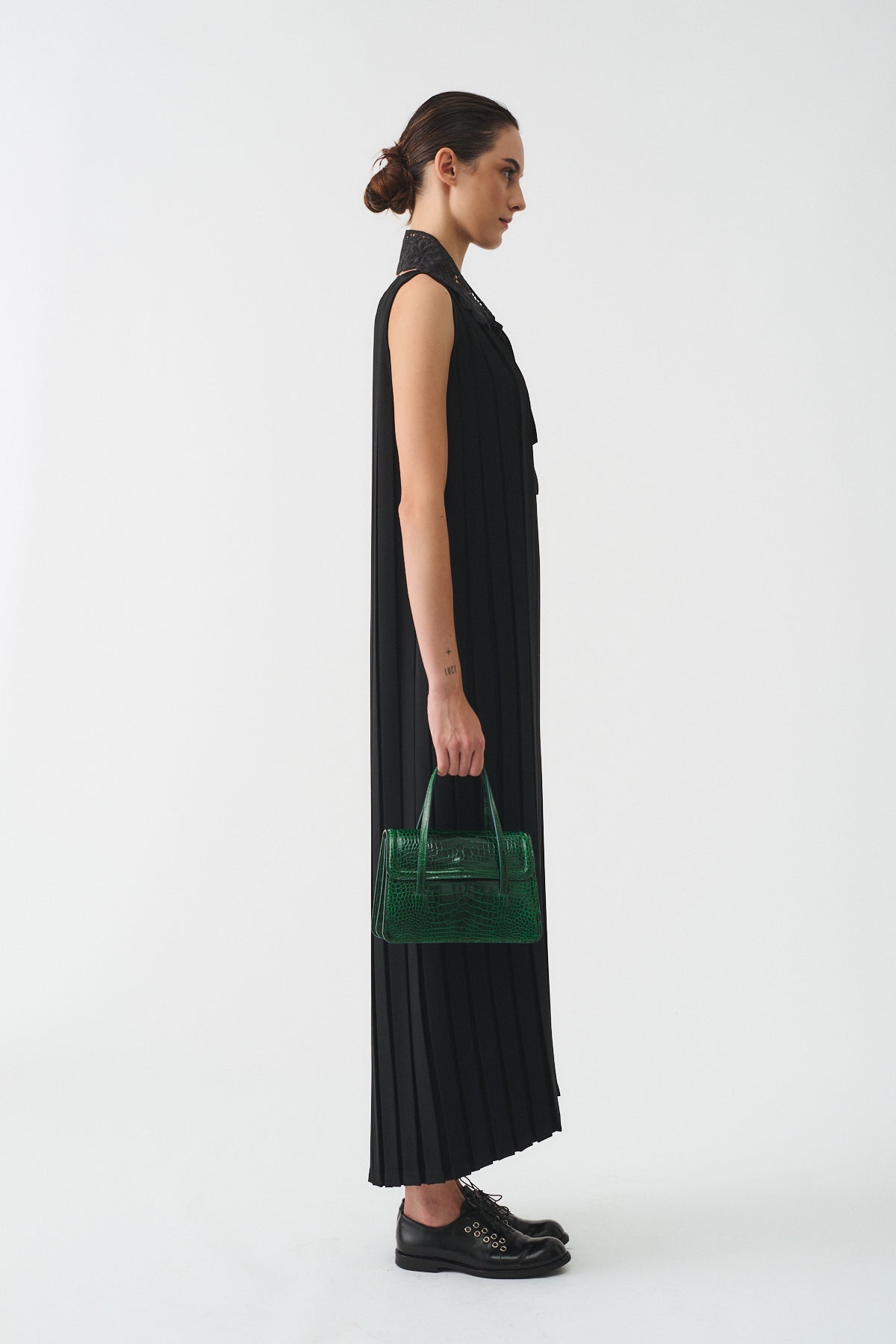 Model wearing a black pleated long dress, holding a green handbag, showcasing elegance and style.