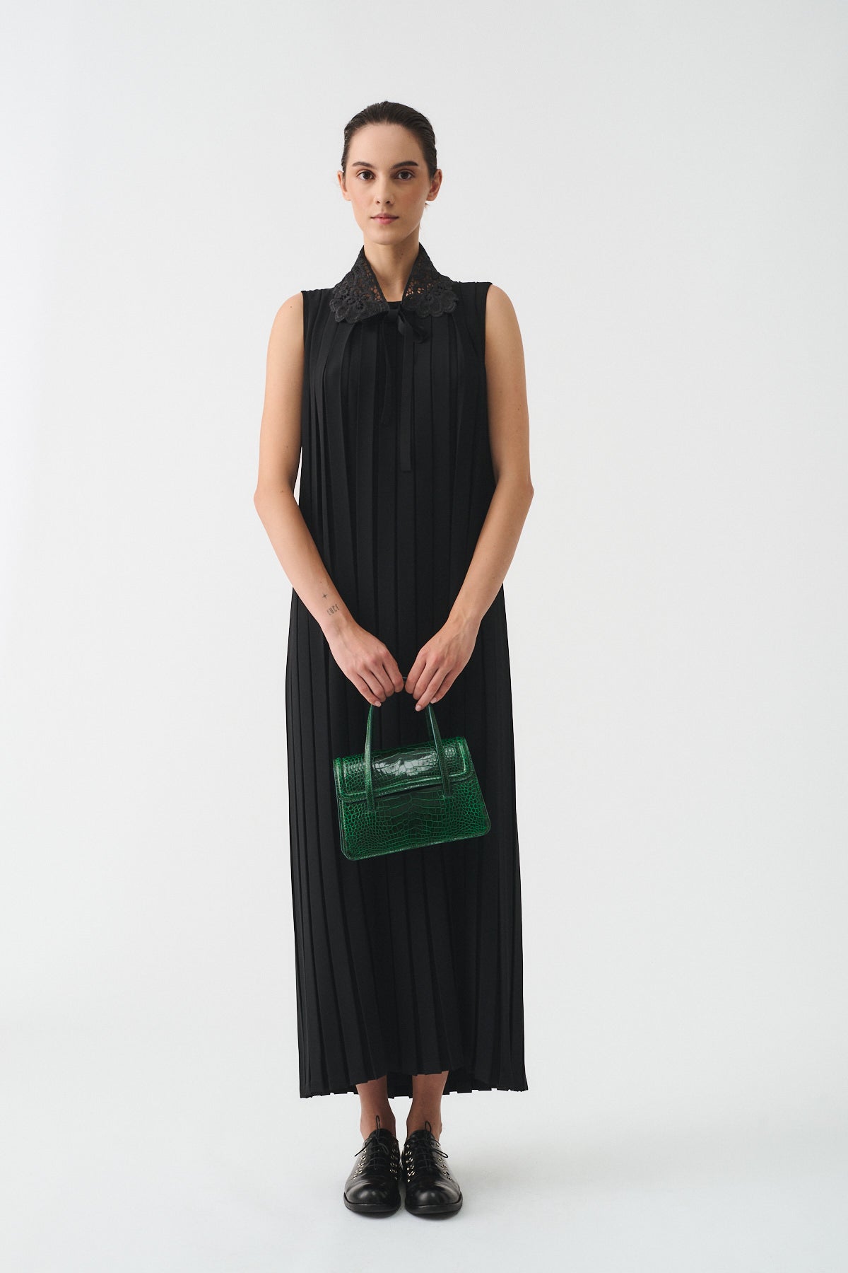 Model wearing a black pleated dress with a collar, holding a green handbag, against a plain background.