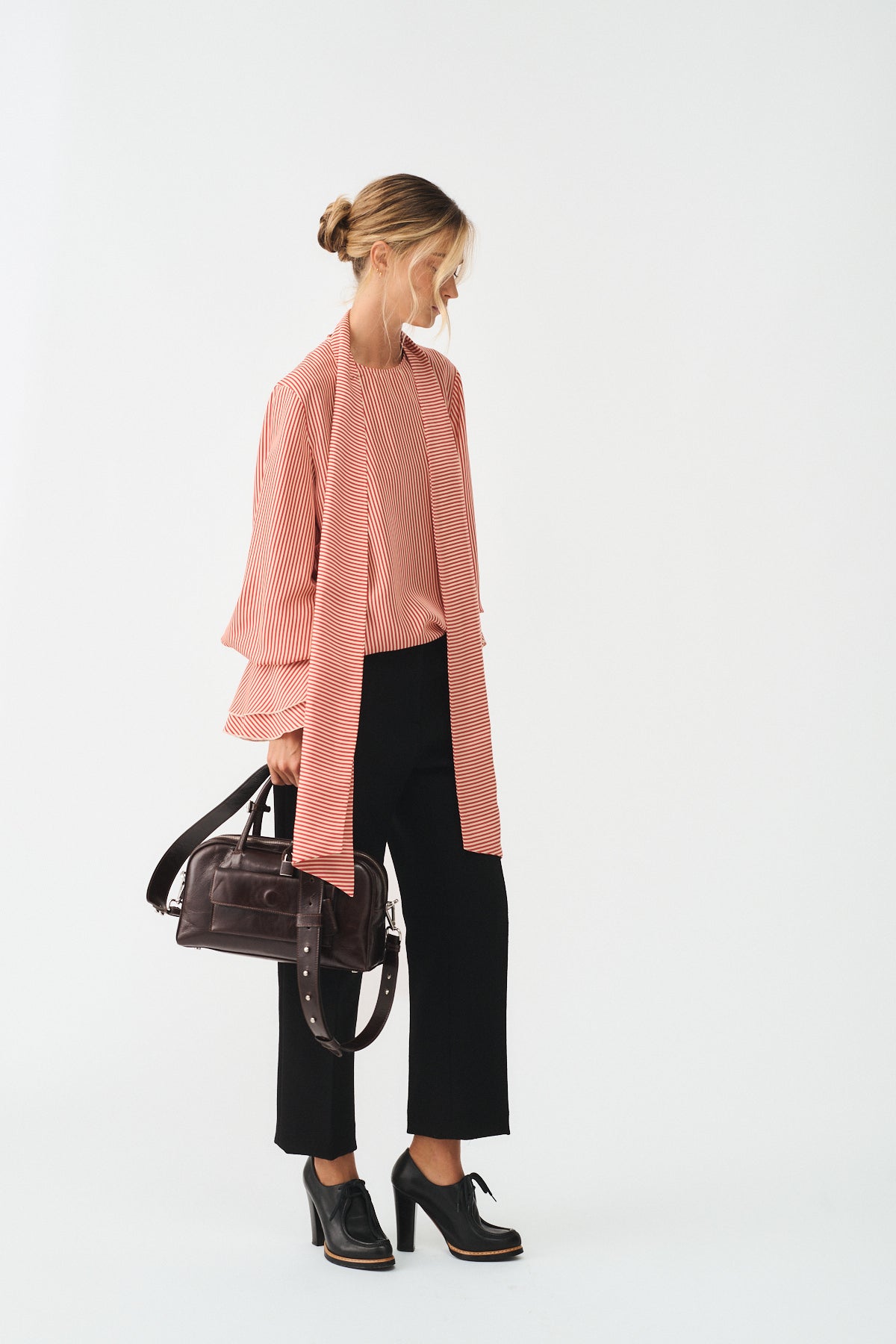 Model wearing a pink cardigan with a matching top, black trousers, and high-heeled shoes, holding a small dark brown handbag.