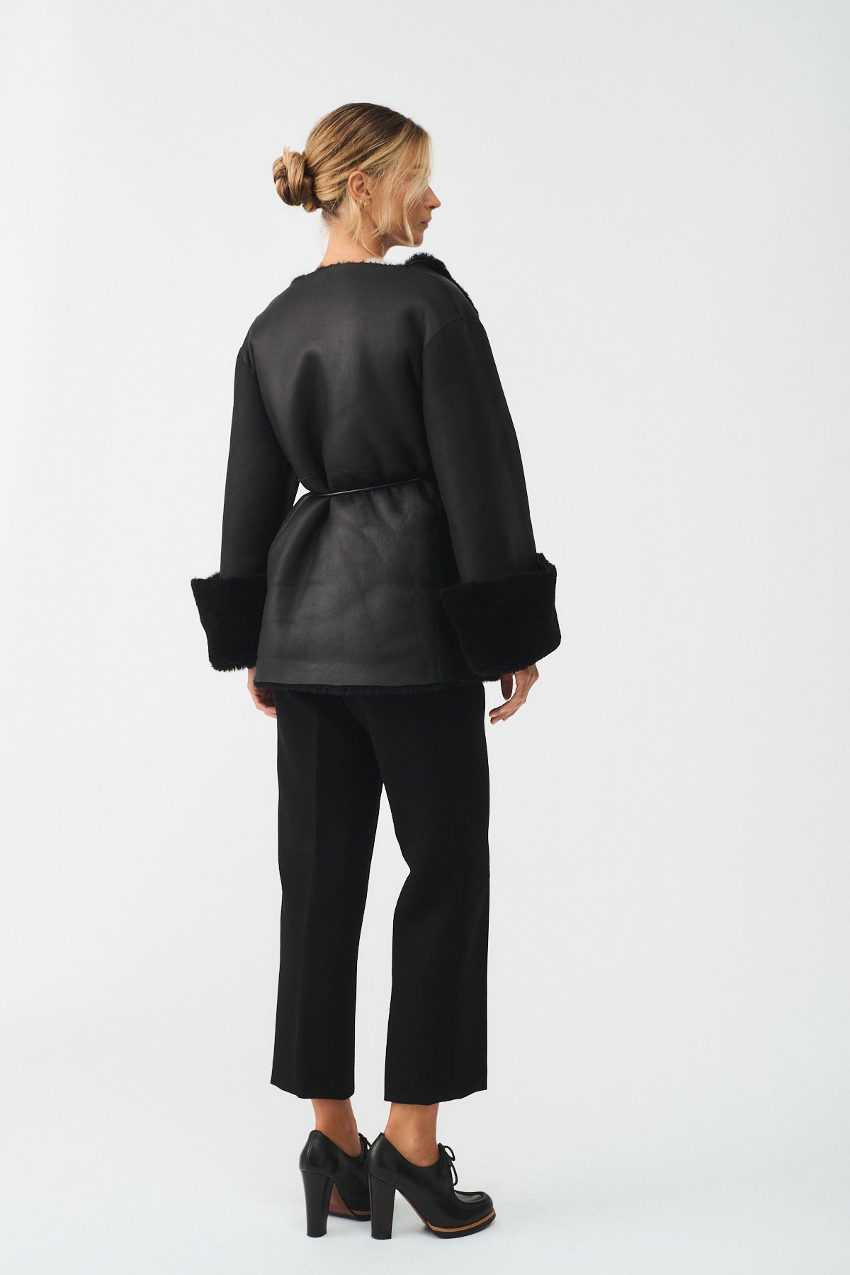 Elegant woman wearing a reversible shearling coat with wide sleeves and a belt, paired with tailored black pants.