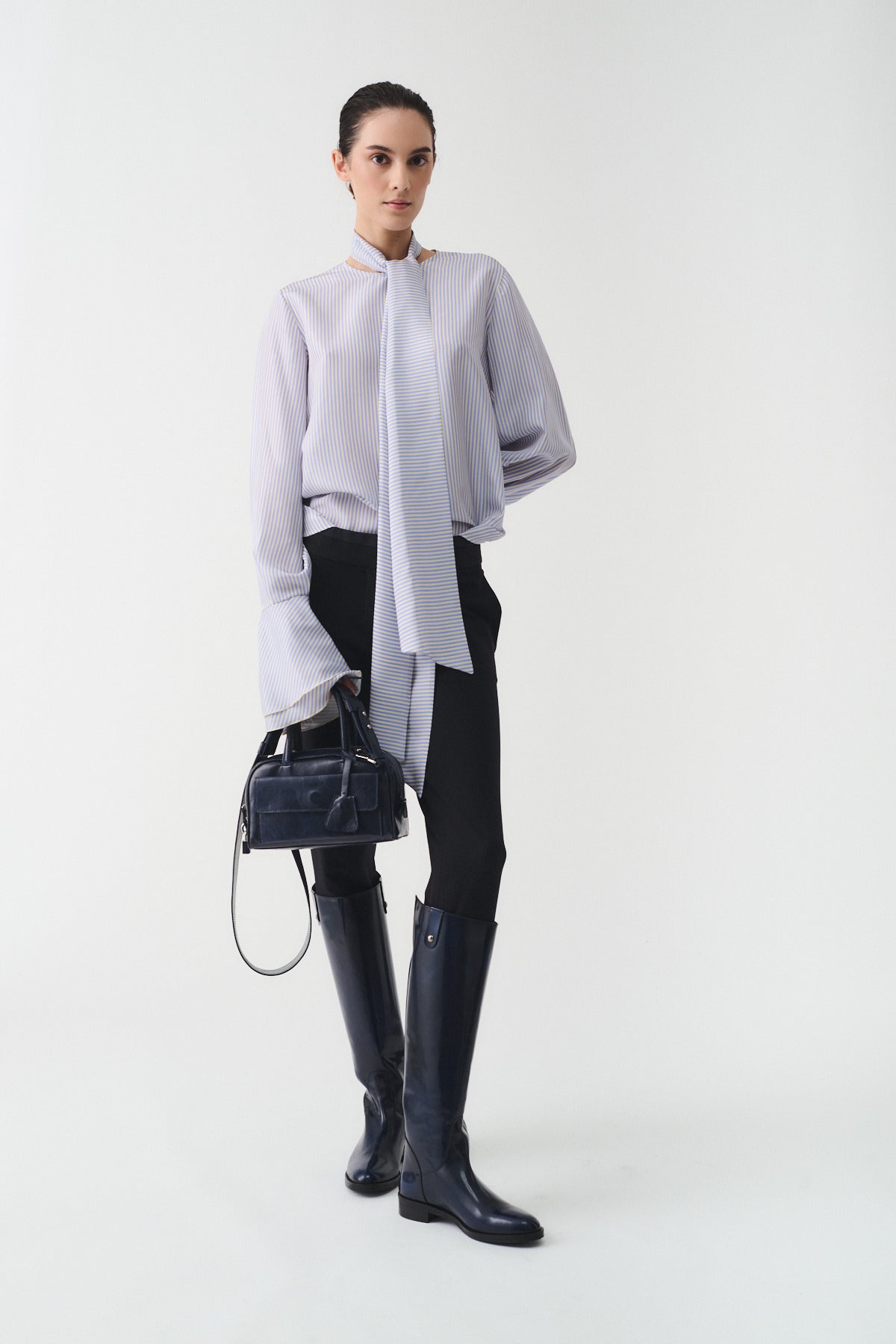 Woman in a stylish outfit featuring a light blouse and black trousers, holding a small handbag and wearing tall boots.