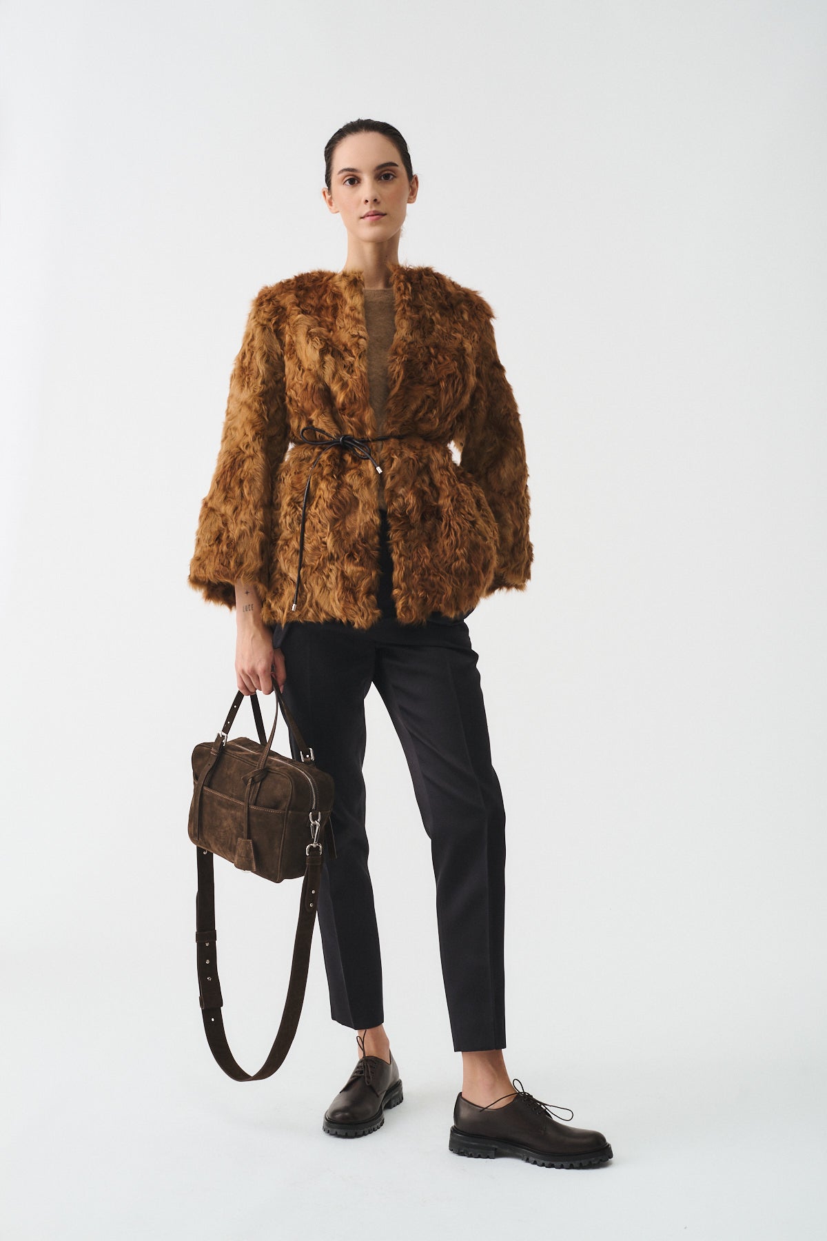 Model wearing a brown faux fur jacket with a belted waist, paired with dark pants and stylish shoes, holding a brown bag.