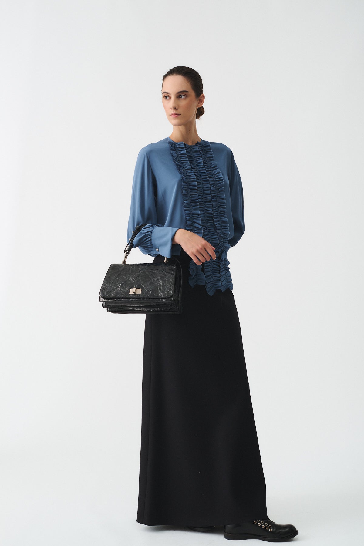 Model wearing a blue ruffled blouse and long black skirt, holding a chic black handbag, showcasing modern elegance.