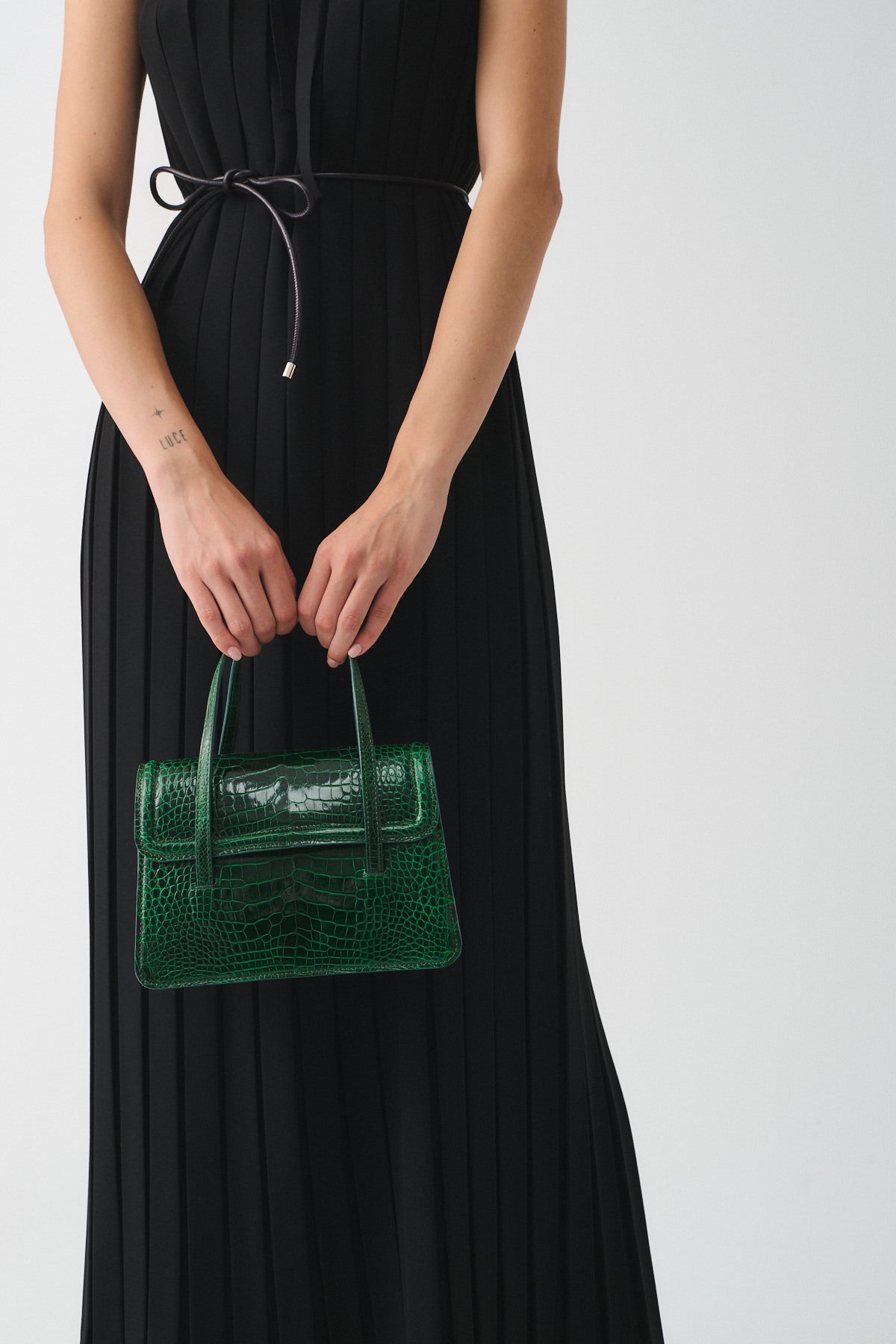 Model holding a stylish green crocodile-effect handbag while wearing a black pleated dress.