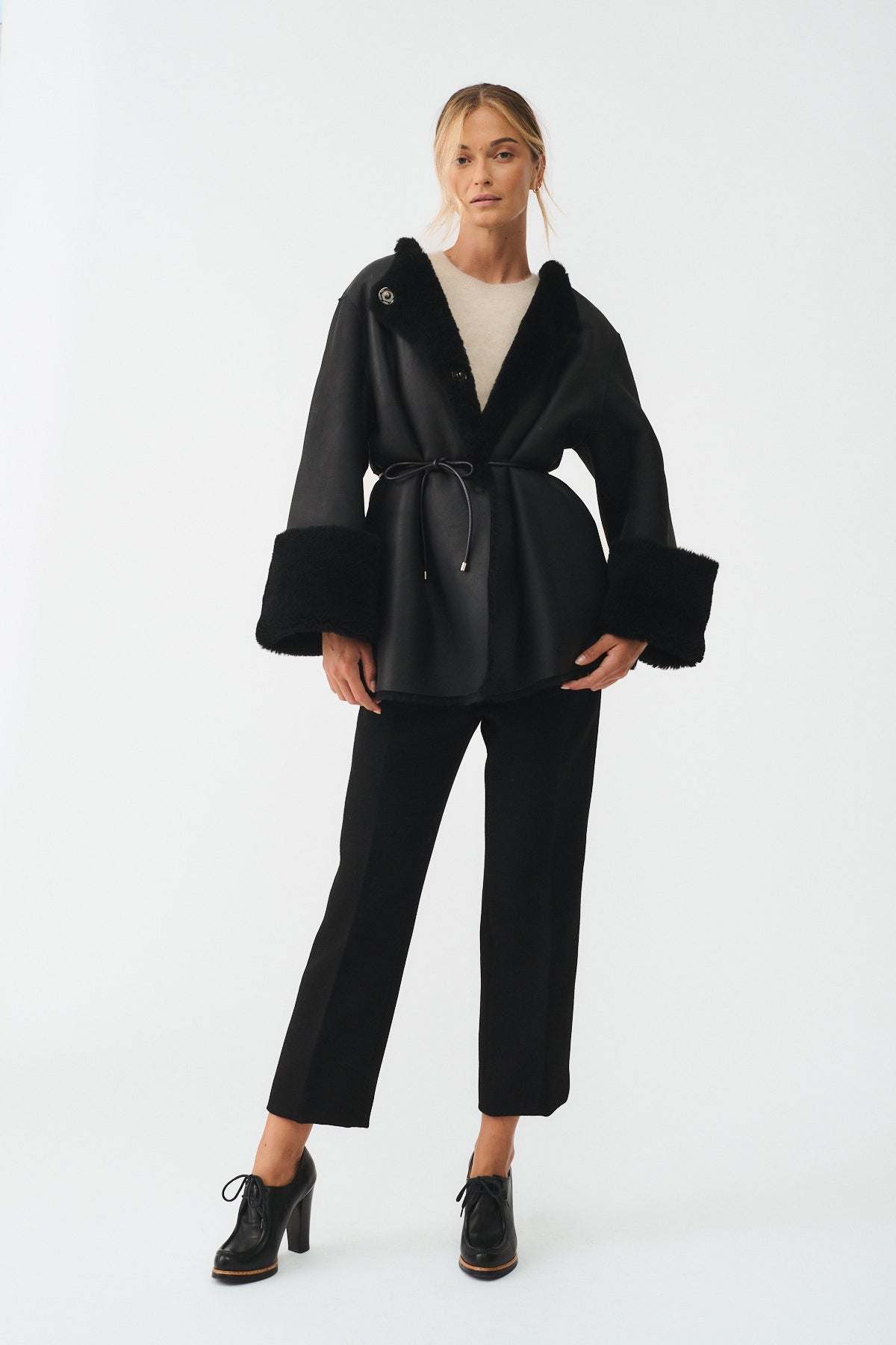 Model wearing a reversible shearling coat with a cinched waist, paired with tailored black pants and heels.