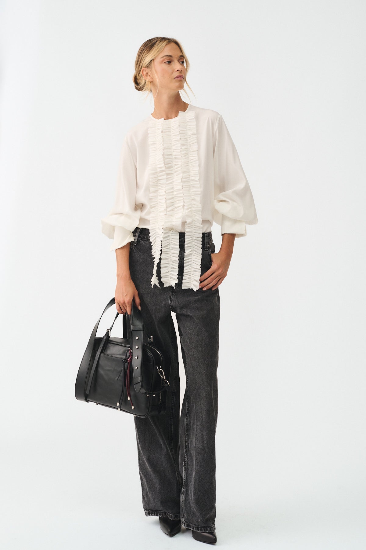 Model wearing a stylish white blouse with ruffles, paired with black jeans and holding a black handbag.